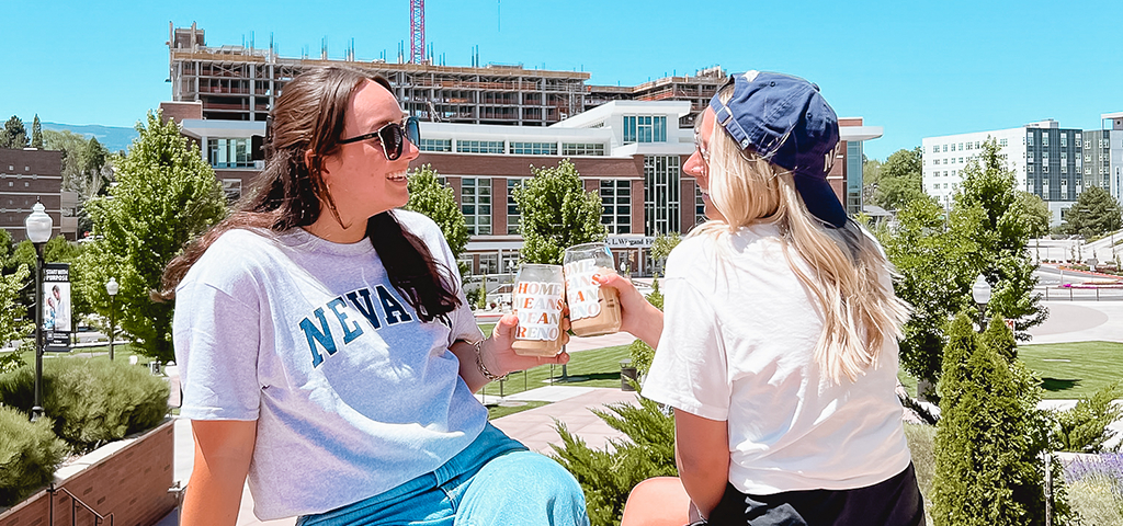 Two young women sitting on the sidewalk in front of a blue wall with 'BATTLE BORN' painted in white