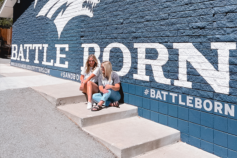 Two young women sitting on the sidewalk in front of a blue wall with 'BATTLE BORN' painted in white